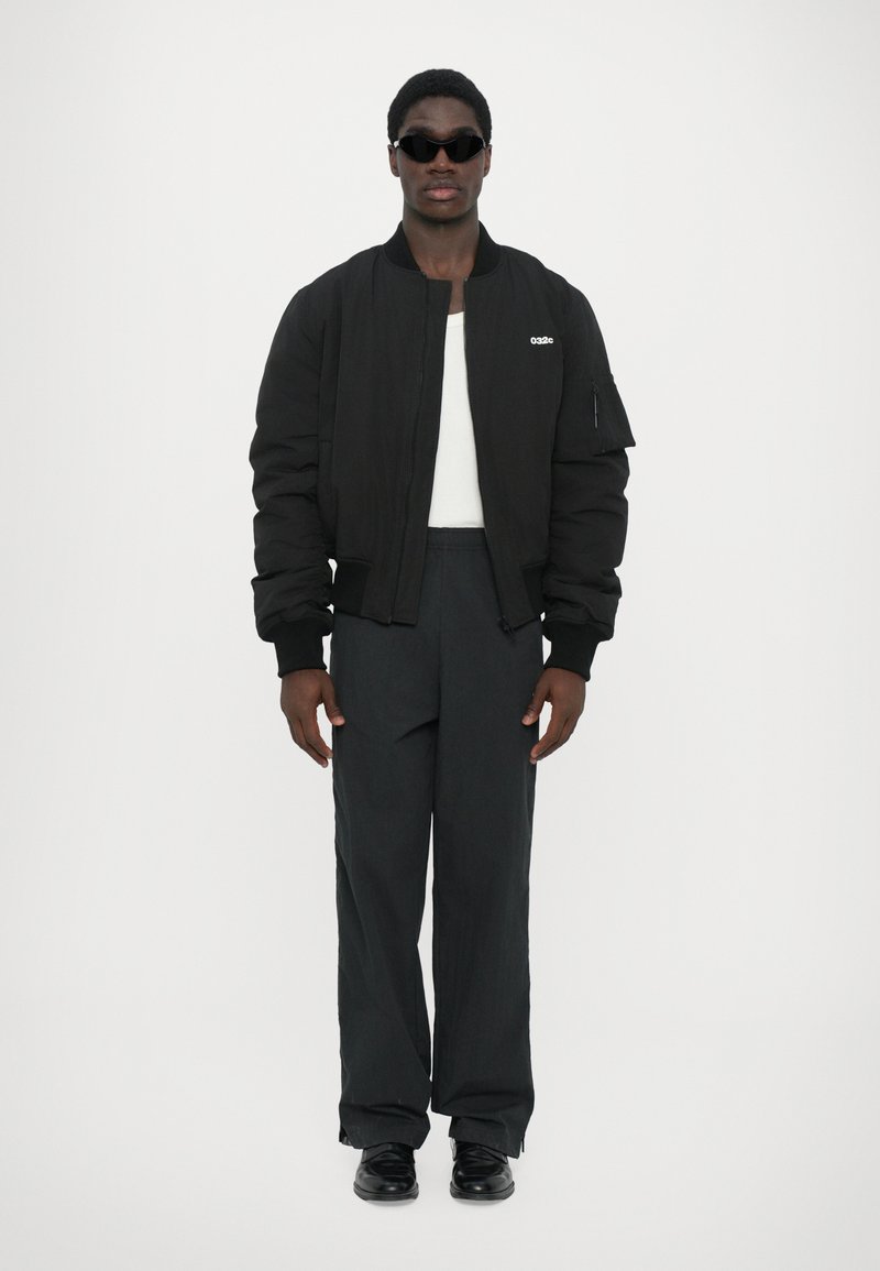 Chaqueta bomber negra con puños acanalados, logo blanco, pantalones negros ajustados y zapatos negros, llevada con gafas de sol negras y una blusa blanca.