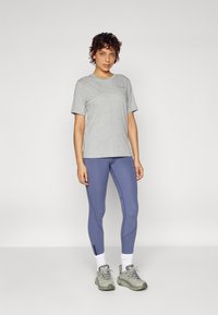 Szürke, rövid ujjú póló, kék leggings, fehér zokni és szürke sportcipő. A leggings oldalvarrásos. Az anyag simának és rugalmasnak tűnik.