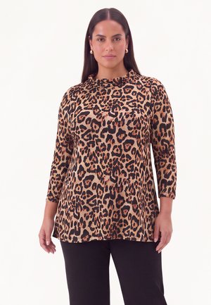 Vrouw met lang donker haar, die gouden oorringen draagt en een bruine lange mouw top met luipaardprint en een zwarte broek, staand voor een witte achtergrond.