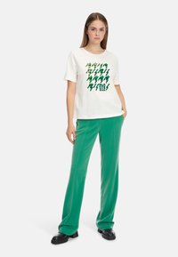 Weißes Baumwoll-T-Shirt mit grünem Blitz-Graphic; kombiniert mit grünen weit geschnittenen Hosen und schwarzen Plateauschuhen. Einfaches Design, lockere Passform.