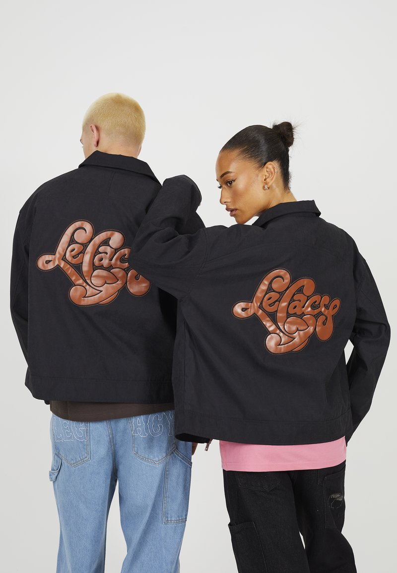 Sorte jakker har et stort brunt læder "Le Casse" logo på ryggen. De parres med lyseblå jeans og en lyserød skjorte nedenunder.