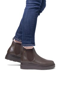 Botas de cuero marrón hasta el tobillo con paneles laterales elásticos, suela elevada y acentos texturizados, combinadas con jeans azul oscuro.