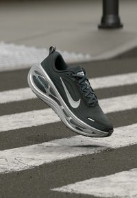 Black and gray athletic shoe with a textured upper, reflective silver swoosh, and a cushioned sole, shown mid-air over a crosswalk.