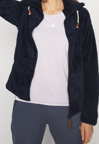 Veste en polaire bleu marine avec une fermeture éclair à l'avant, un col haute et des accents à cordon de serrage. Elle présente une texture douce et un design ajusté, portée sur un haut léger.