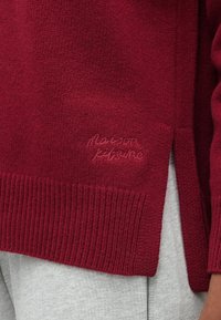 Burgundy knit sweater with ribbed hem and cuffs. Features a subtle embroidered logo on the side and a side slit for ease of movement.