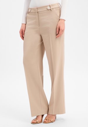 Pantalon beige à jambes larges avec détails de boutons à la taille, porté avec des talons nus ouverts et un haut blanc à manches longues, modèle montré de la taille vers le bas.