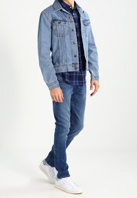 Light blue denim jacket with two front pockets, paired with a navy and black plaid shirt, blue jeans, and white sneakers.