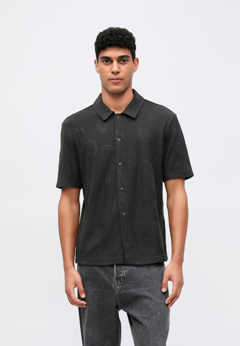 Svart kortermet polo-skjorte med en subtil teksturert mønster, med knappestolpe foran og en avslappet passform. Kombinert med mørke grå jeans.