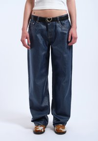 Breda mörkblå denimbyxor med en glänsande finish, utrustade med ett svart bälte med ett hjärtformat spänne och synliga sömnadsdetaljer.