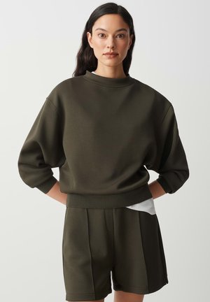 Donna con capelli scuri indossa una felpa larga verde oliva e pantaloncini abbinati, mani dietro la schiena su uno sfondo chiaro uniforme.