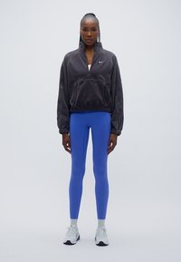 Πουλόβερ φλις σε σκούρο γκρι με φερμουάρ και μπροστινή τσέπη, συνδυασμένο με μπλε κολάν και λευκά αθλητικά παπούτσια με γκρι σόλες.