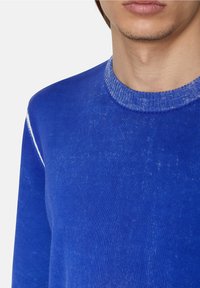 Blå stickad tröja med rund halsringning, med en texturerad yta och vita accentstygn längs axelsömmarna.