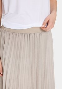 Gonna plissettata beige con vita elasticizzata, caratterizzata da una texture liscia. Abbinata a una maglietta bianca, le mani sistemano il tessuto in vita.