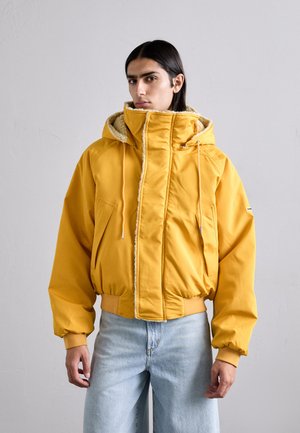 Yellow puffer jacket with a hood, featuring a fuzzy lining, side pockets, and ribbed cuffs. Paired with light blue jeans.