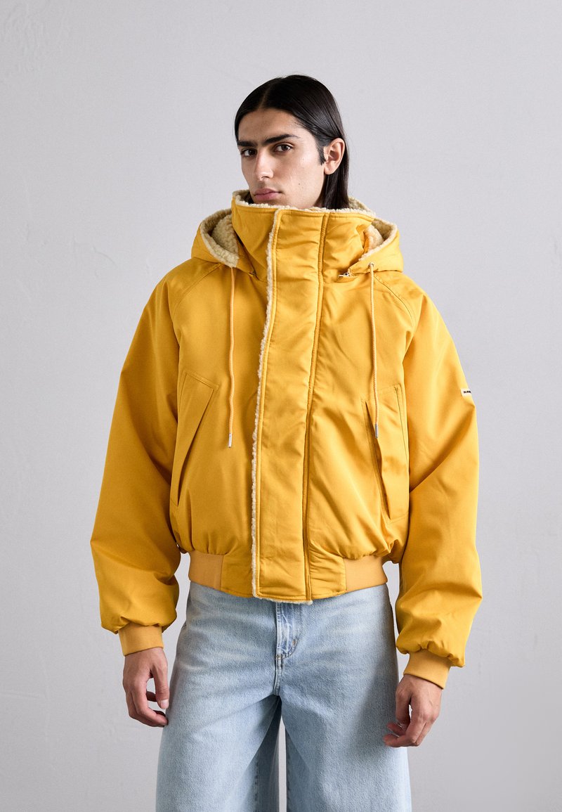 Yellow puffer jacket with a hood, featuring a fuzzy lining, side pockets, and ribbed cuffs. Paired with light blue jeans.
