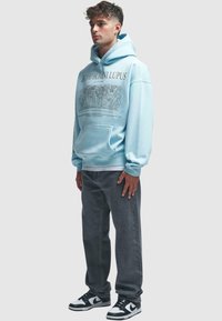 Light blue hoodie with a graphic print on the front, paired with gray denim pants and black and white sneakers. Hooded design, front pocket.