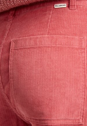 Vaaleanpunaiset corduroy-housut, joissa on pystysuorat poimut ja takatasku, jossa on etiketti. Kankaassa on pehmeä tekstuuri ja näkyviä ompeluyksityiskohtia.