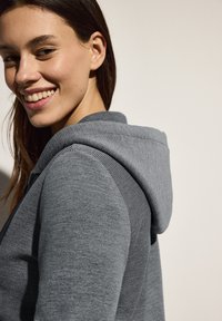 Felpa grigia a strisce con un tessuto testurizzato, dotata di cappuccio, cuciture visibili e una vestibilità slim. Il modello sorride, mettendo in risalto il design dell'indumento.