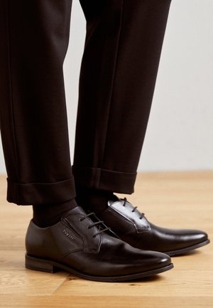 Black leather dress shoes with a smooth finish, lace-up design, and a slight pointed toe, paired with cropped black trousers.