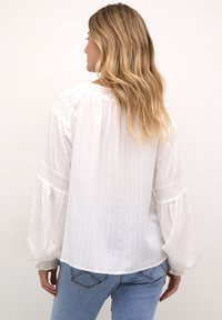 Vit blus med långa, puffiga ärmar, texturerade ränder och elastiska detaljer vid ärmsluten, tillsammans med blåjeans.