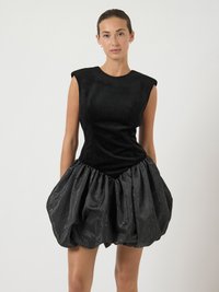 Mulher vestindo um vestido preto sem mangas, com corpete estruturado e saia de bolha texturizada e volumosa, em pé contra um fundo claro e liso.