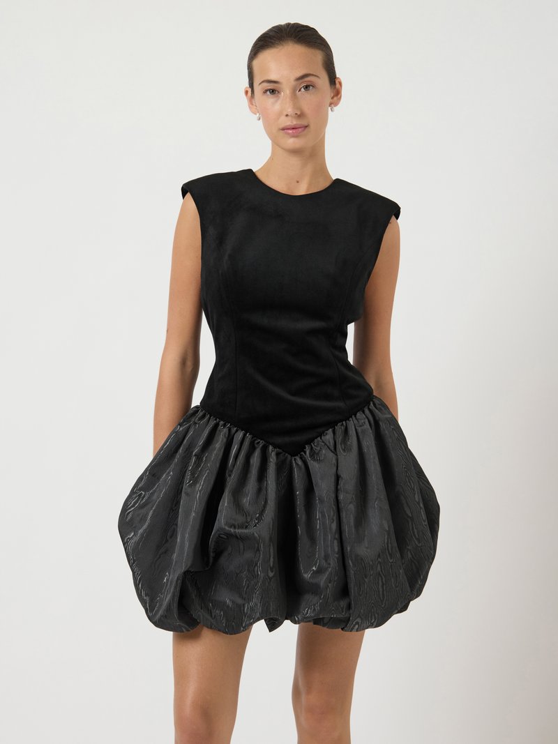 Mulher vestindo um vestido preto sem mangas, com corpete estruturado e saia de bolha texturizada e volumosa, em pé contra um fundo claro e liso.