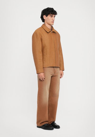 Abrigo marrón con textura, con cuello, cierre frontal y bolsillos laterales, combinado con pantalones marrones a juego y zapatos negros. Diseño simple, tonos neutros.