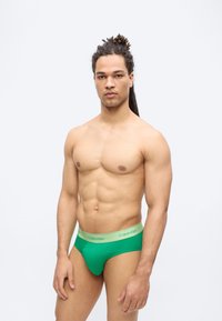 Μυώδης άνδρας με μακριά dreadlocks φορώντας πράσινα εσώρουχα Calvin Klein, στέκεται μπροστά σε απλό λευκό φόντο.