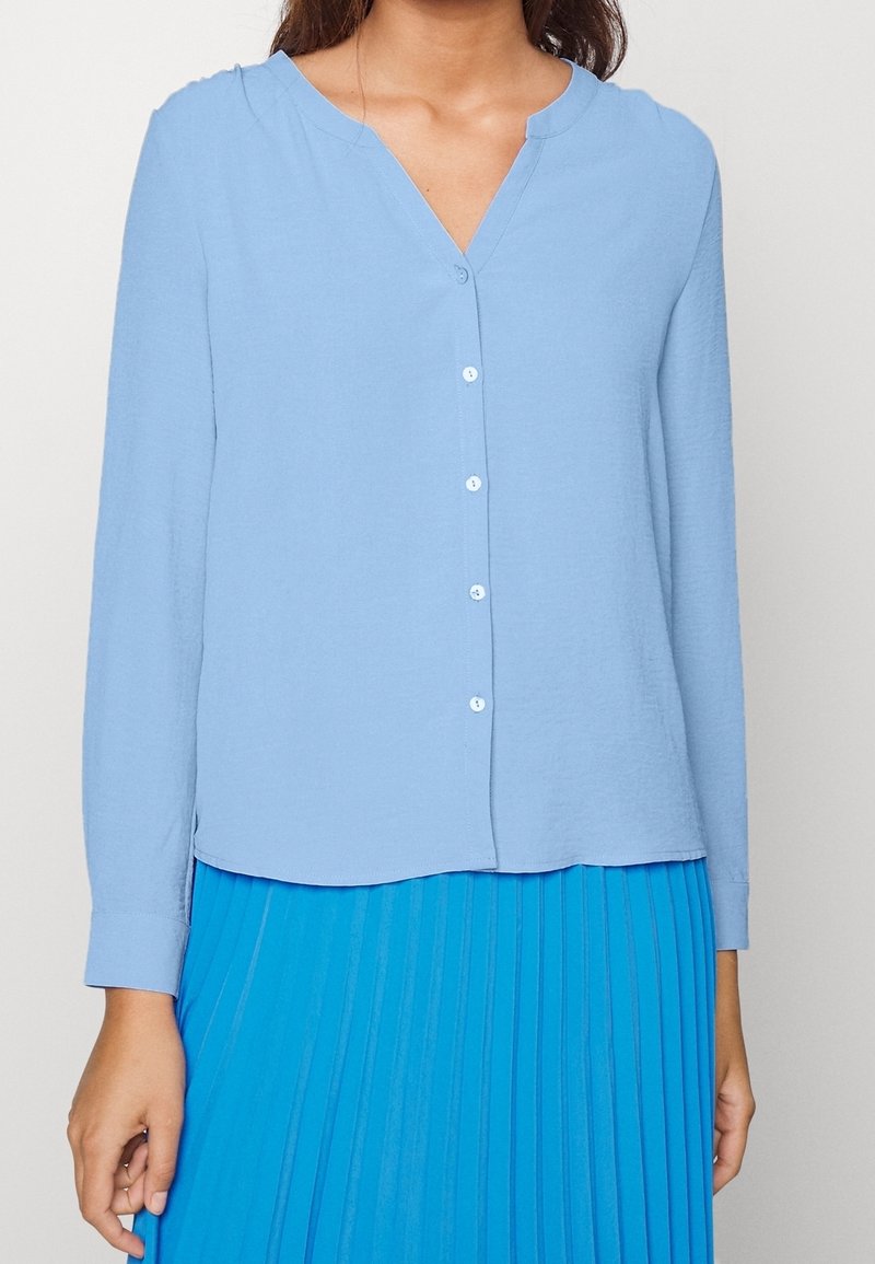 Femme portant un chemisier bleu clair à manches longues boutonné et une jupe plissée bleu vif sur un fond uni.