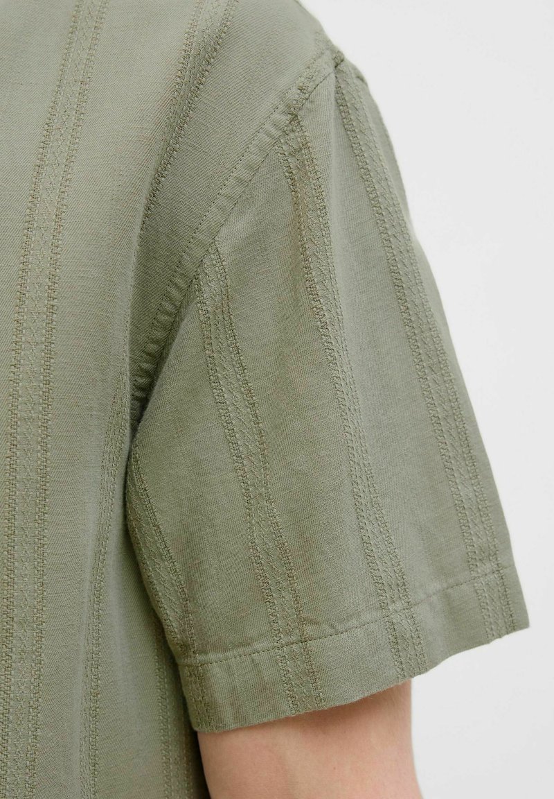Camisa de manga corta en algodón verde oliva con rayas texturizadas. Presenta costuras dobles a lo largo de las costuras y un corte sencillo y relajado.