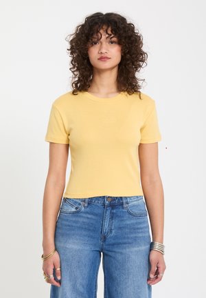 Jeune femme aux cheveux bouclés portant un haut court jaune à manches courtes et un jean taille haute bleu, debout devant un fond blanc uni.