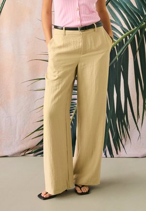 Osoba ubrana w beżowe spodnie o szerokich nogawkach z czarnym paskiem, różowy prążkowany top bez rękawów i czarne sandały z kwadratowym noskiem, stojąca na tle tropikalnych roślin.