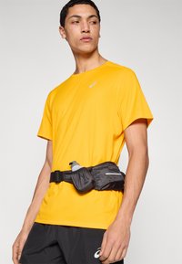 Κίτρινη μπλούζα με αδιάβροχη ιδιότητα, κοντά μανίκια, σε συνδυασμό με μαύρη τσάντα μέσης που περιέχει μπουκάλι νερού, με ανακλαστική ταινία.