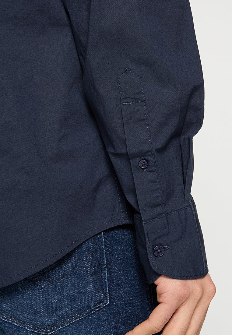 Chemise à manches longues bleu marine avec poignets boutonnés, coupe ajustée, et texture en coton lisse. Dotée d'un col pointu et de détails de couture subtils.