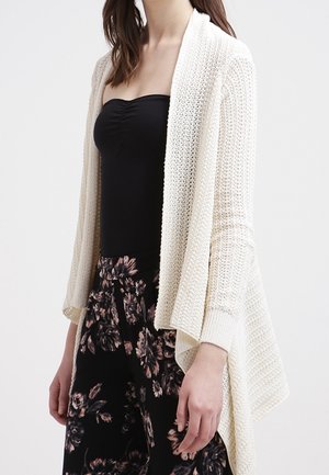 Kvinde iført en draperet cremet strikcardigan over en sort stropløs top og sorte blomstrede bukser med vide ben.
