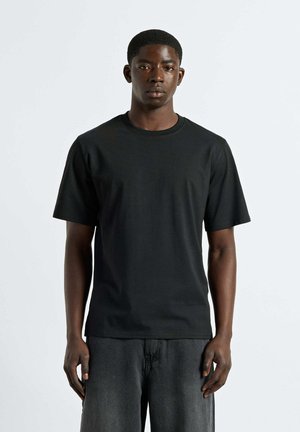 Young man with short hair wearing a plain black short-sleeve t-shirt and dark gray pants standing against a light background.