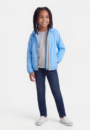 Enfant souriant aux cheveux tressés, portant une veste bleu clair, un t-shirt gris, un jean foncé et des chaussures blanches sans lacets, debout avec une main dans la poche.