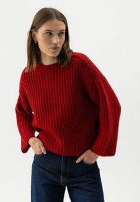 Una donna che indossa un maglione rosso spesso e jeans blu scuro, in piedi con le mani in tasca, guarda verso sinistra su uno sfondo neutro.