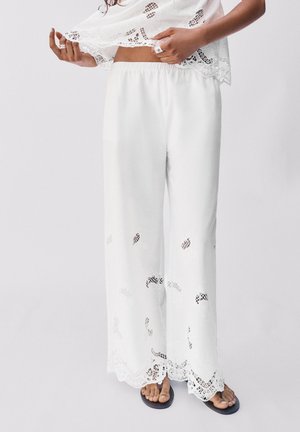 Vrouw draagt witte wijde broek met kanten uitsnijdingen en geschulpte zomen, gecombineerd met zwarte slippers op een effen achtergrond.