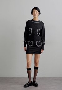 Fekete kötött pulóver és szoknya szett fehér vonalakkal díszített zsebekkel, hosszú ujjakkal és gombos záródással. Kiegészítők közé tartozik a combig érő zokni és a loaferek.