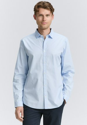 Young man with light brown hair wearing a light blue button-up shirt and dark trousers, standing against a plain light background.