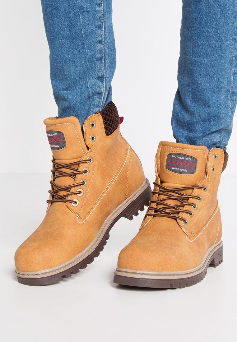 Bottes de travail marron clair à lacets avec col rembourré portées avec un jean bleu sur fond blanc.