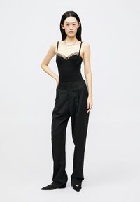 Mustmust mustrase lace detailidega mustas, koos vaba musta pükste komplektiga. Modelil on sirged juuksed ja ta seisab ühtlase tausta ees.