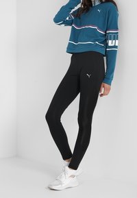 Teal kort sweatshirt med vita och lila linjer, i kombination med svarta leggings. Modellen har på sig vita sneakers. Mjuk tygkvalitet och figurnära design.