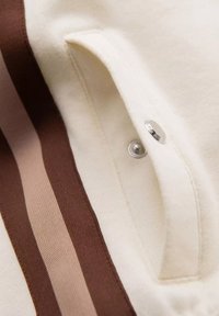 Tissu couleur crème avec des rayures marron et beige, comportant une fermeture à bouton-pression et un petit détail de poche. Texture douce.