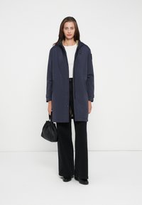 Cappotto blu navy con cappuccio e chiusura a bottone, progettato con foderato morbido e tasche laterali, abbinato a pantaloni neri a gamba larga e una borsa nera.