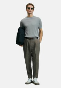 Grijze geribbelde gebreide t-shirt, lichtgrijze broek, zwarte riem, houdt een donkere jas vast, zonnebril en witte sneakers met groene accenten.