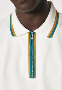 Polo blanc avec un col zippé, ornée de rayures turquoise et jaunes. Tissu doux, design épuré et détail de fermeture éclair métallique.