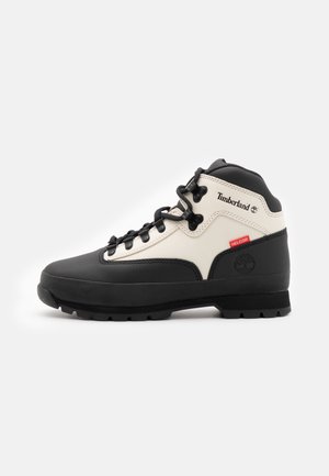 Wysoki trekkingowy but Timberland w czarno-białym kolorze z czarnymi sznurówkami, solidną podeszwą oraz czerwoną metką z materiału Helcor na boku.