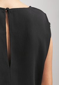 Svart topp med rund hals, korta ärmar och ett nyckelhål bak, gjord av ett slätt tyg med en något matt textur.